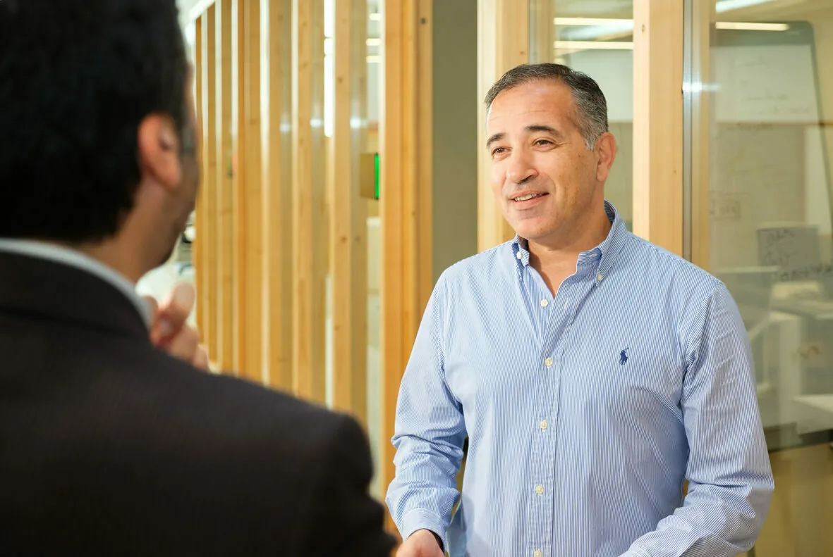 A man in a blue button-up shirt talks to another man in a suit in an office hallway with wooden dividers.