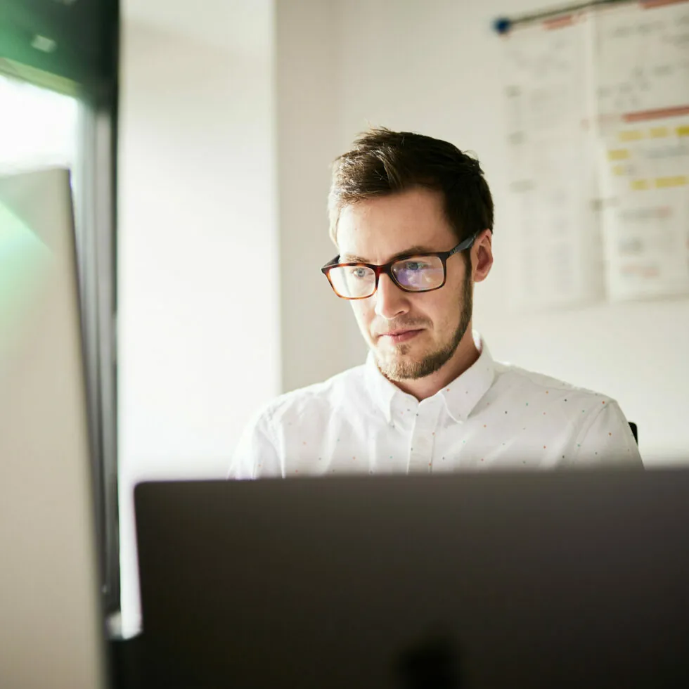 A man, wearing glasses and a white shirt with small polka dots, is focused on his work while using a laptop in a brightly lit office. A calendar is visible on the wall behind him.