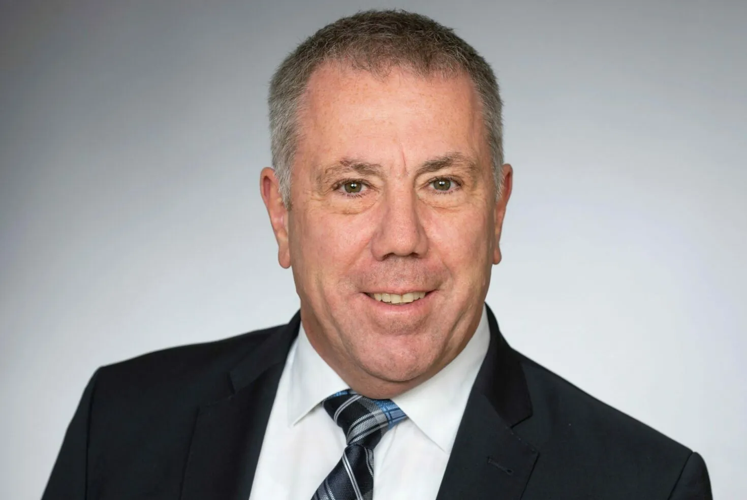 A middle-aged man with short gray hair wearing a dark suit, white shirt, and striped tie, smiling against a plain gray background.