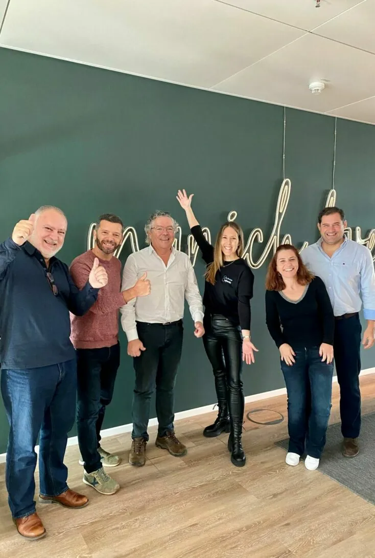 A group of six people, five men and one woman, posing in front of a wall with neon signage. Some are smiling and giving thumbs up, while the woman raises her arm and smiles.