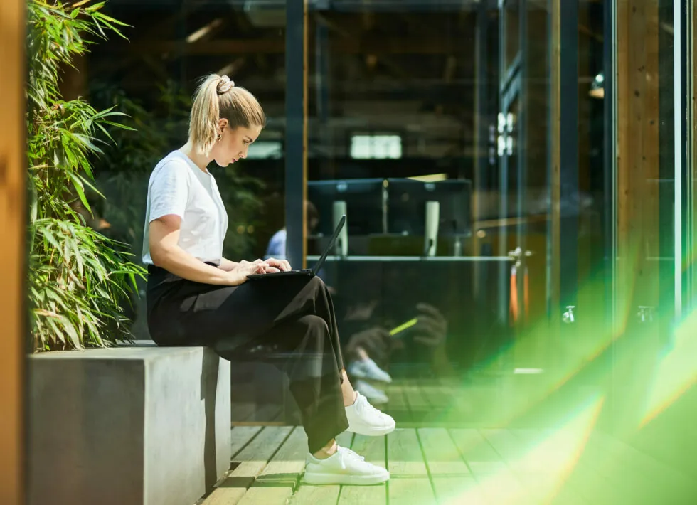 A person with a ponytail, dressed in a white shirt and dark pants, sits on an outdoor bench working on a laptop. The setting includes plants and glass windows reflecting indoor office space.