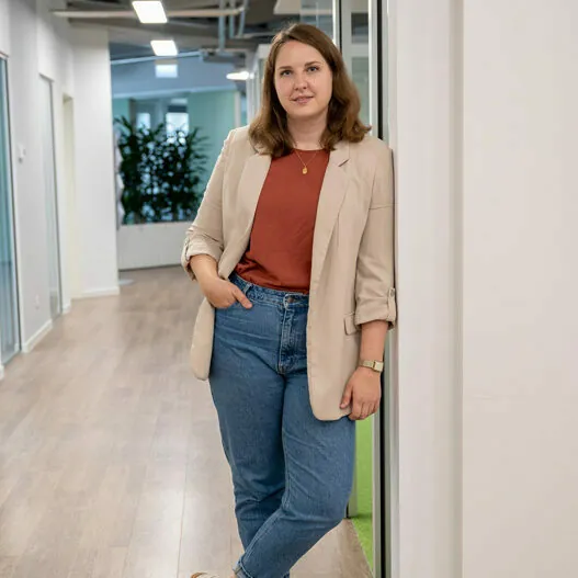 A person is leaning against a wall in a hallway wearing a beige blazer, rust-colored shirt, and blue jeans. The hallway has wooden flooring and plants in the background.