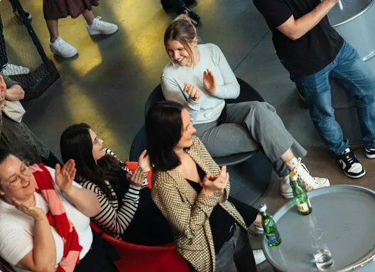 A group of people sitting and standing in a casual setting, some are clapping and smiling. Beverages are placed on a nearby surface.