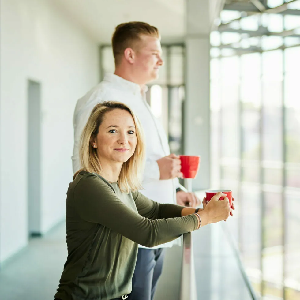 Two people stand by a window holding red mugs, with one person facing the camera and the other looking outside.