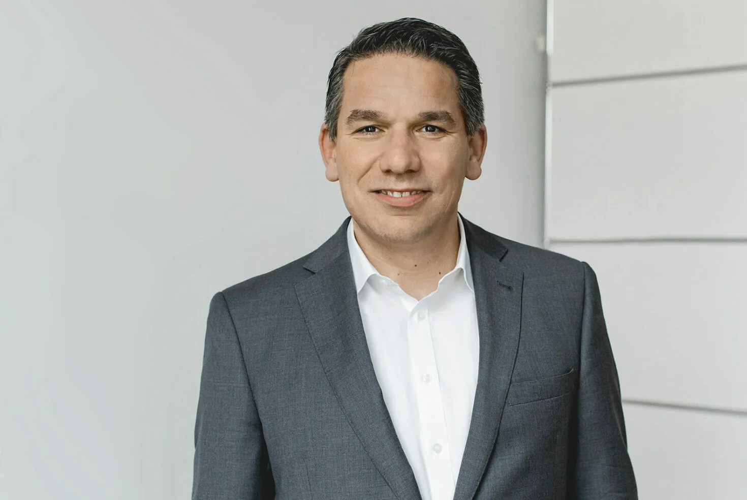 A man wearing a grey suit and white shirt stands against a plain background while looking into the camera and smiling.