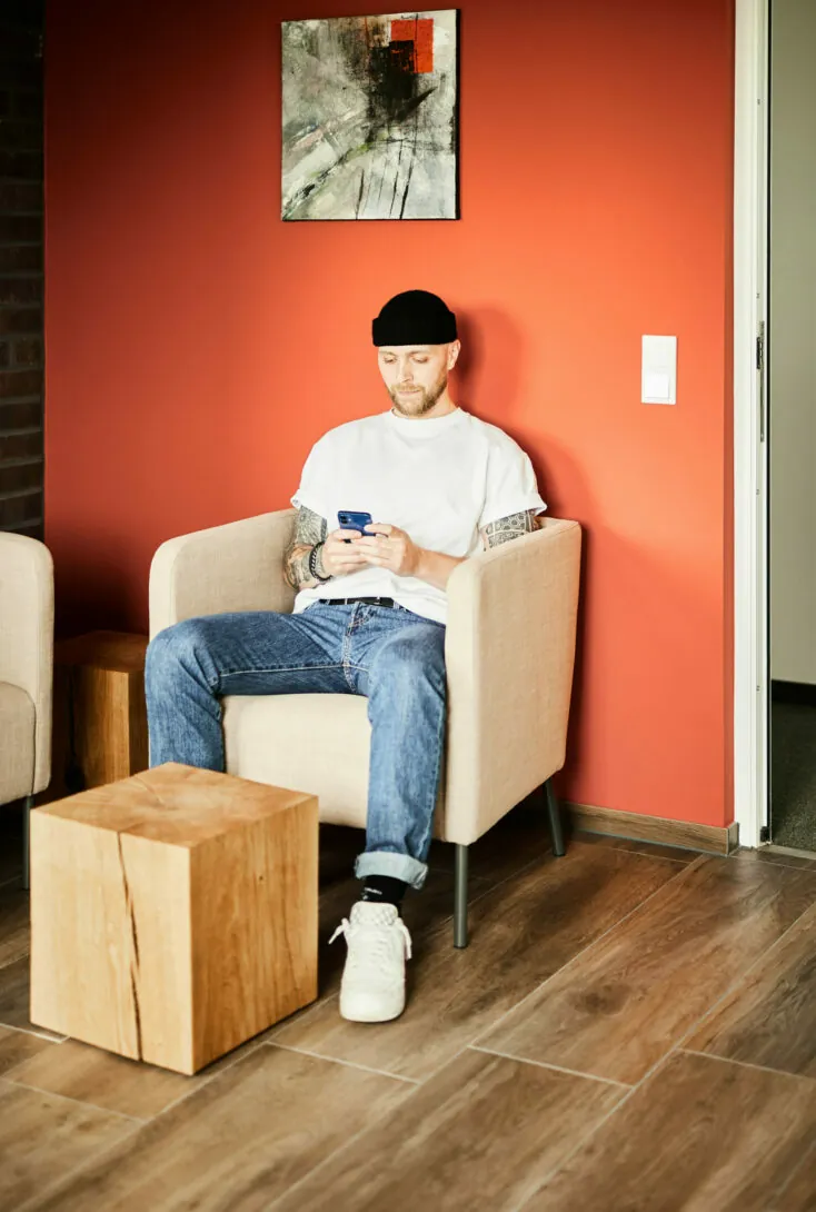 A man with a beard and tattoos sits on a beige chair against a red wall, using a smartphone. He is wearing a black beanie, white T-shirt, blue jeans, and white shoes. There is a wooden cube-table nearby.