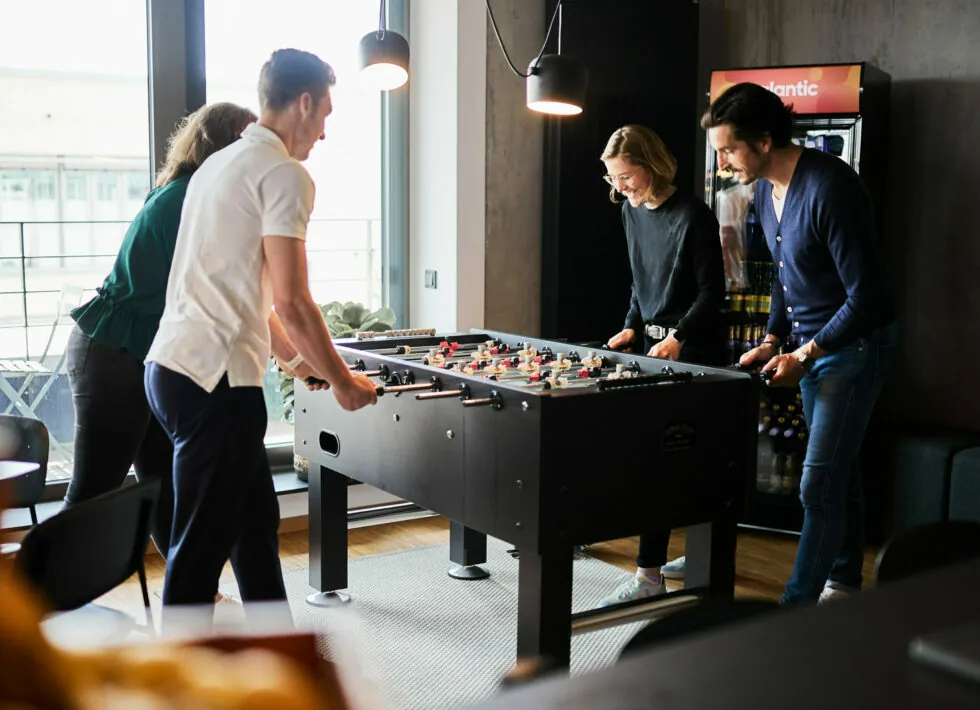 Four people are engaged in a game of foosball in a modern indoor setting. A vending machine and large windows are visible in the background.