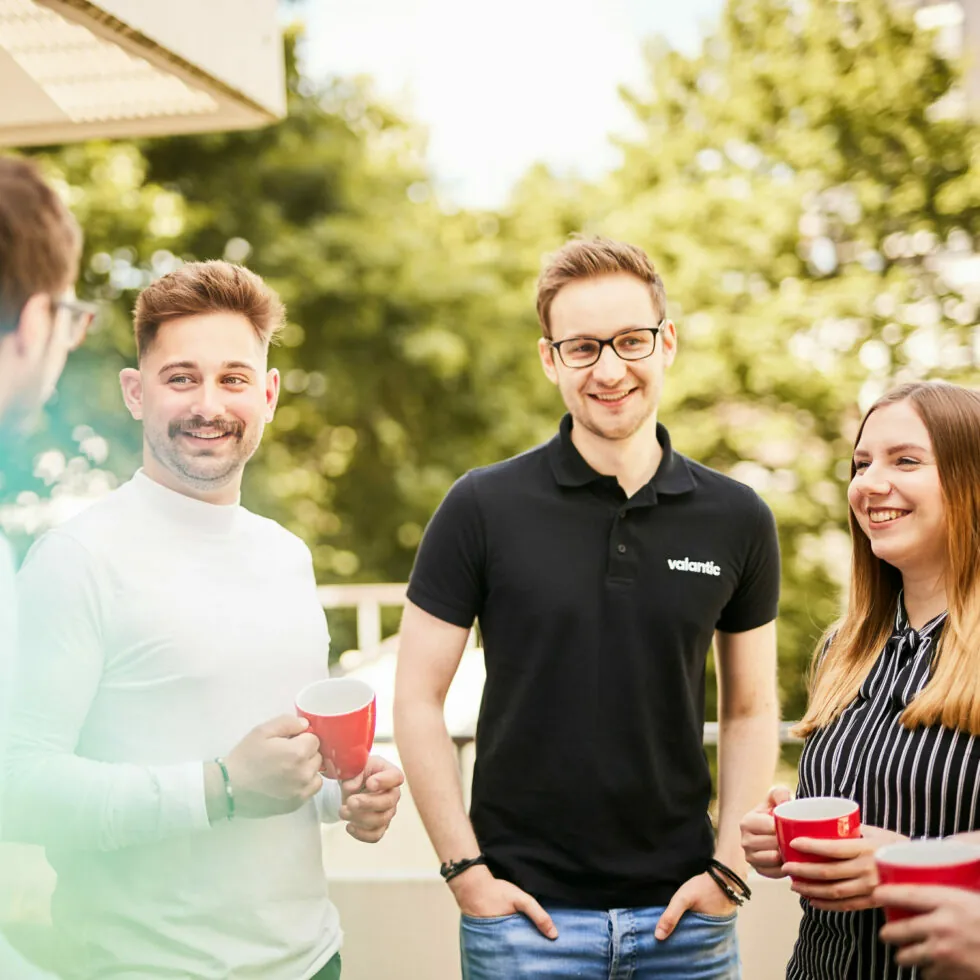Four people are standing outside, smiling and holding coffee mugs. Trees and a building are visible in the background.
