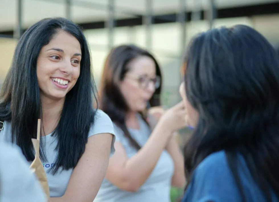 A woman with long black hair smiles and converses with another person while holding a beverage. A second woman in the background, wearing glasses, is drinking. The setting appears to be outdoors.