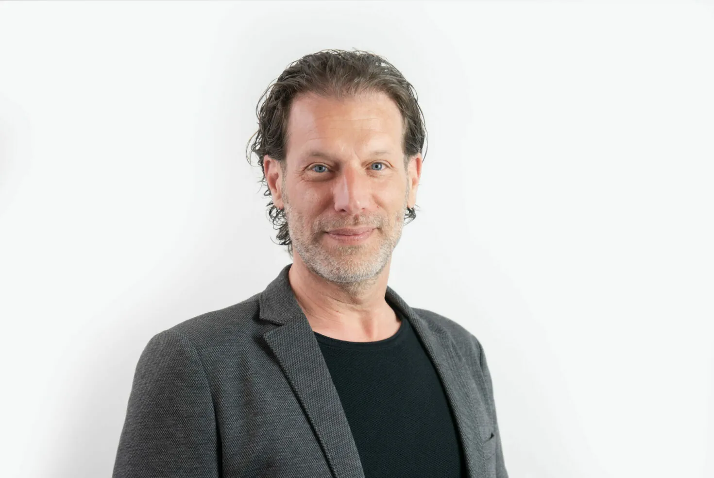 A man with short, slightly curly hair and a beard is wearing a gray blazer and black shirt, posing in front of a plain white background.
