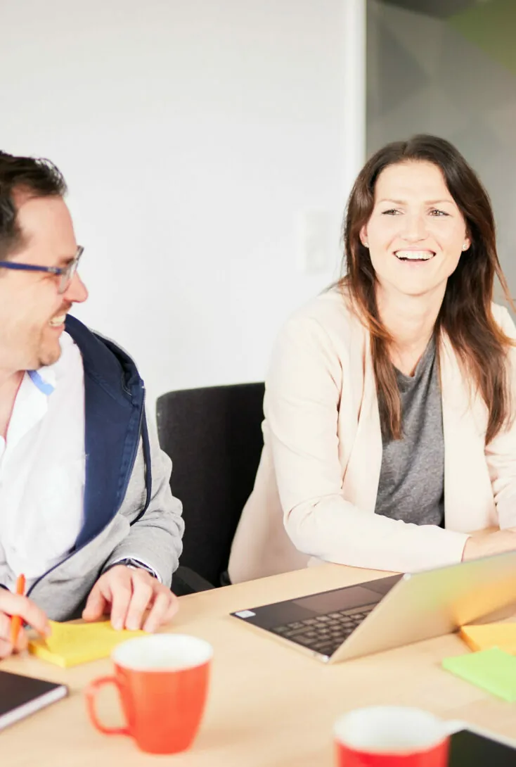 Two people sit at a table laughing, with laptops and coffee mugs in front of them.