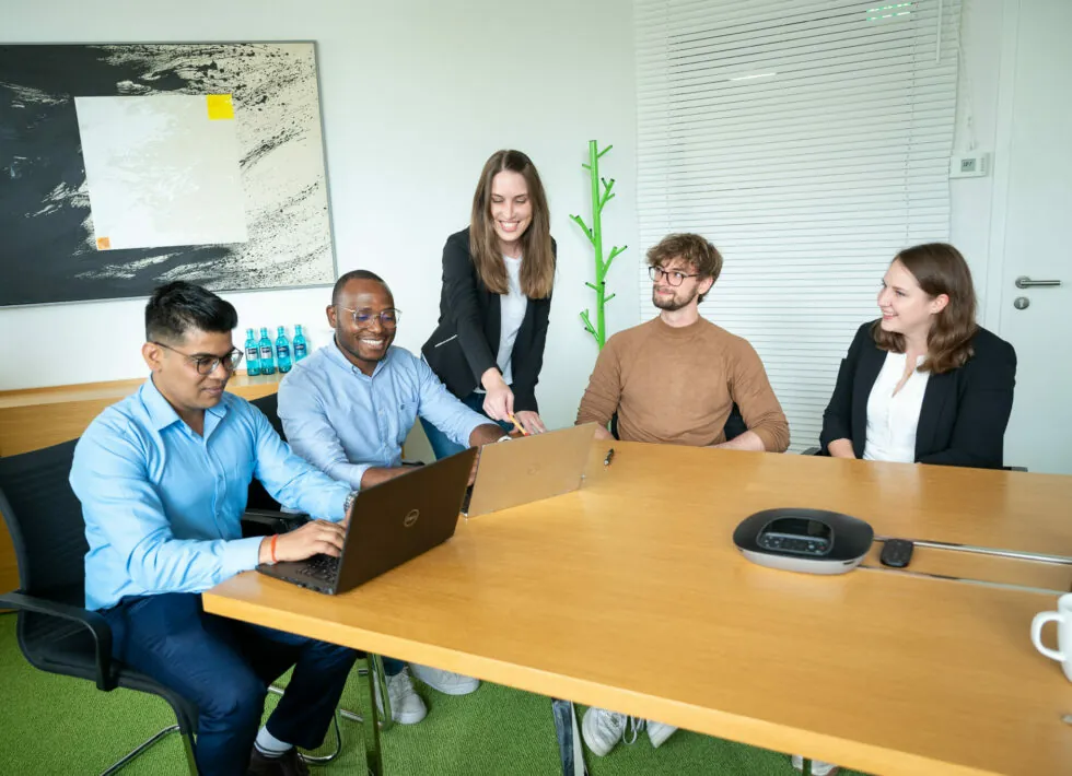 A group of five people sit and stand around a conference table with laptops, engaged in a discussion, in a modern office setting.