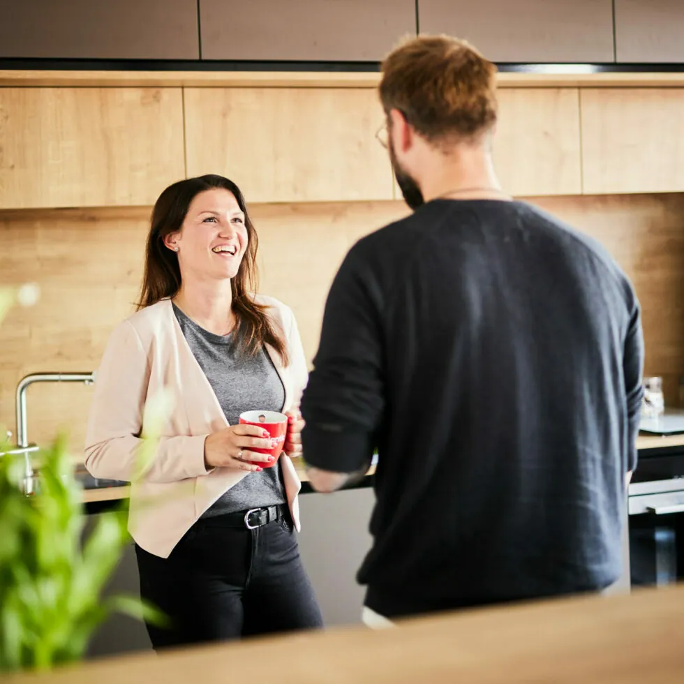 A woman and a man stand in a modern kitchen, conversing. The woman is holding a red mug, smiling, while the man is facing away, partially blurred. There is greenery in the foreground.