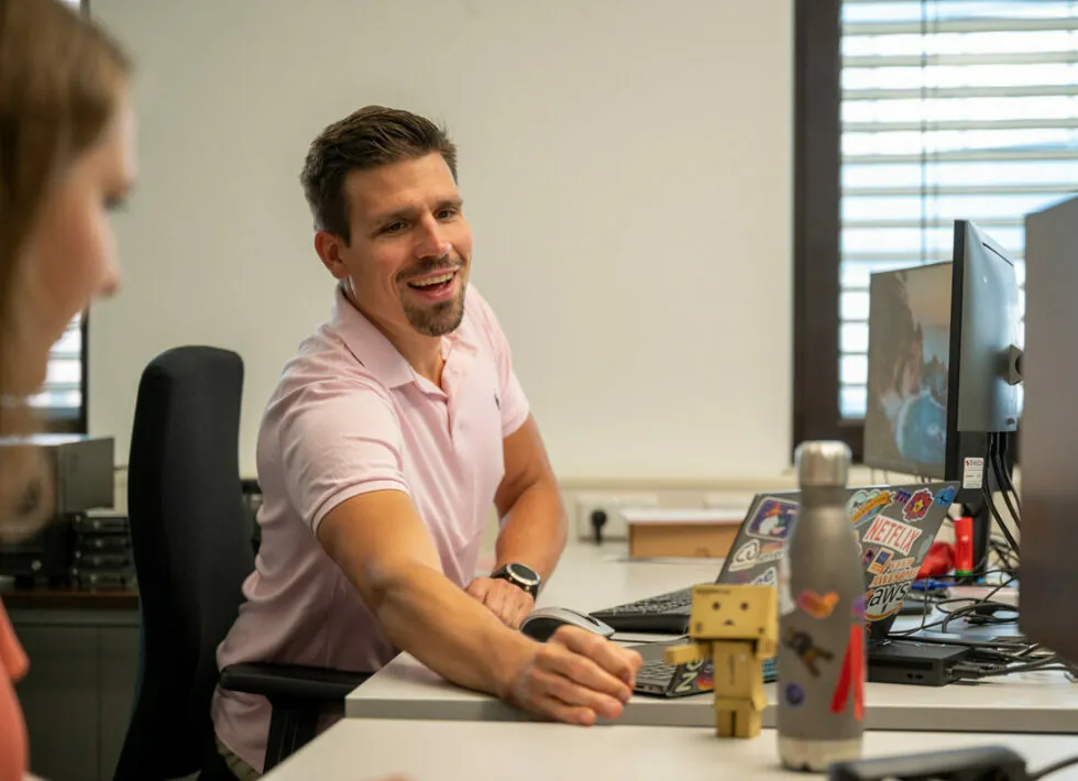 Man in a pink shirt smiling and pointing at a small cardboard robot figurine on a desk in an office setting, with two monitors and various items around. Another person is sitting beside him.