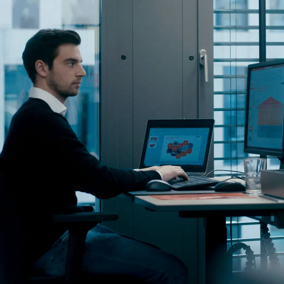 A man works at a desk with a laptop and a monitor in an office setting. Both screens display graphs and charts. The office has large windows with blinds.