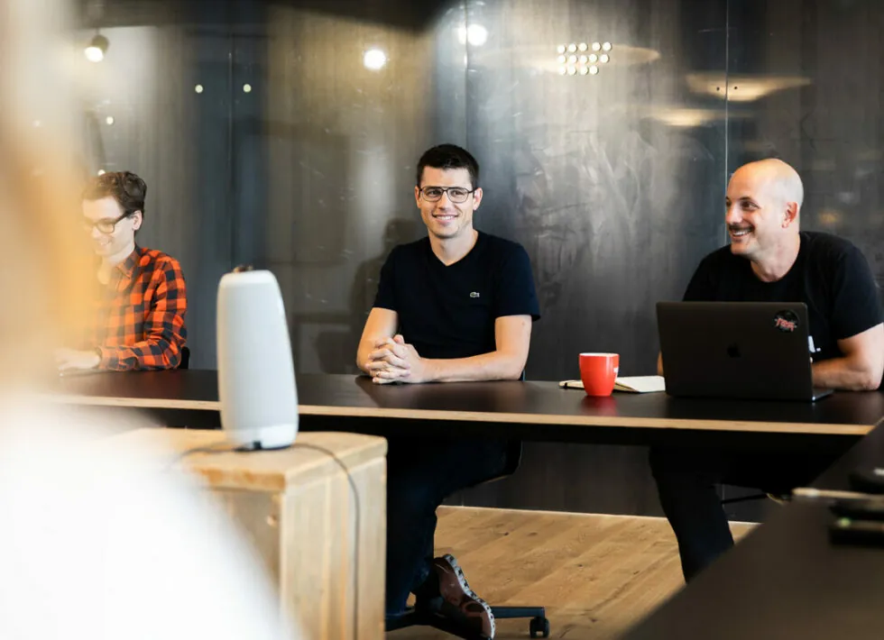 Three people are seated at a conference table, engaged in a meeting. Two men in the center and right are facing the camera, while another person is partially visible on the left.