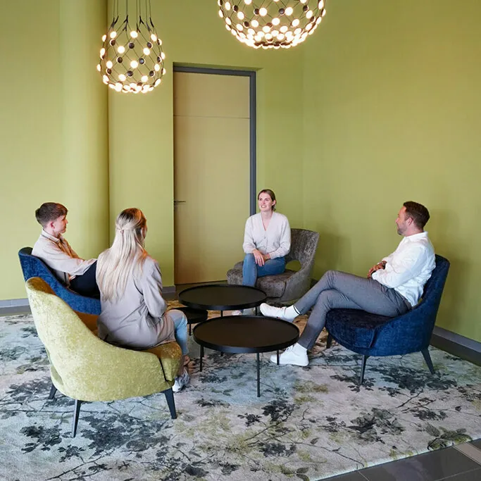 Four people are sitting on colorful chairs around black round tables in a brightly lit room with green walls and hanging lights.
