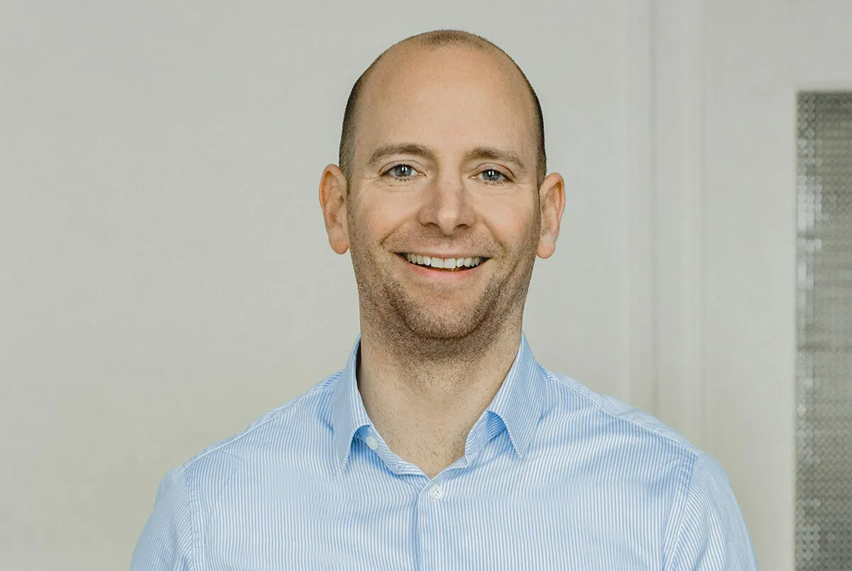 A man with a light complexion, wearing a light blue collared shirt, stands against a plain background, smiling.