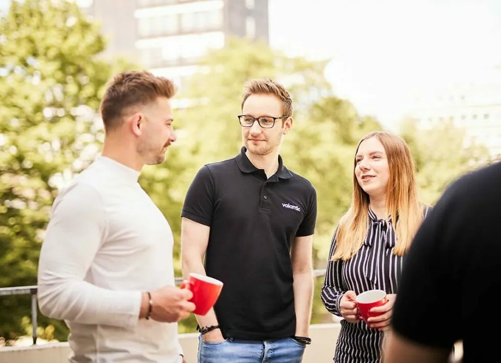 Three people standing outdoors, holding red mugs, and conversing. Trees and buildings are visible in the background.