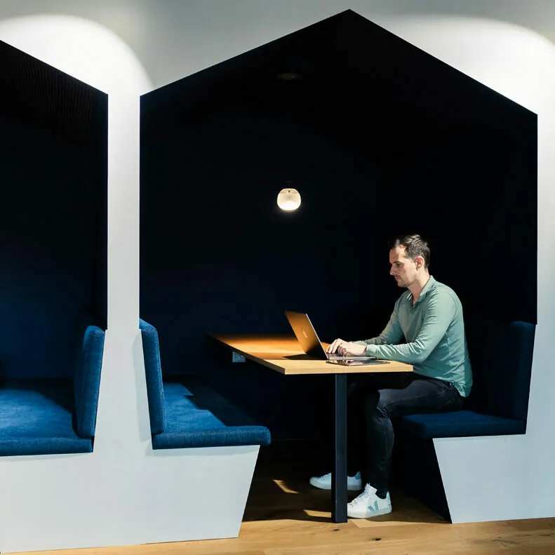 A person is seated at a table inside a modern, minimalist booth, working on a laptop. The booth has dark, cushioned seating and a small overhead light.