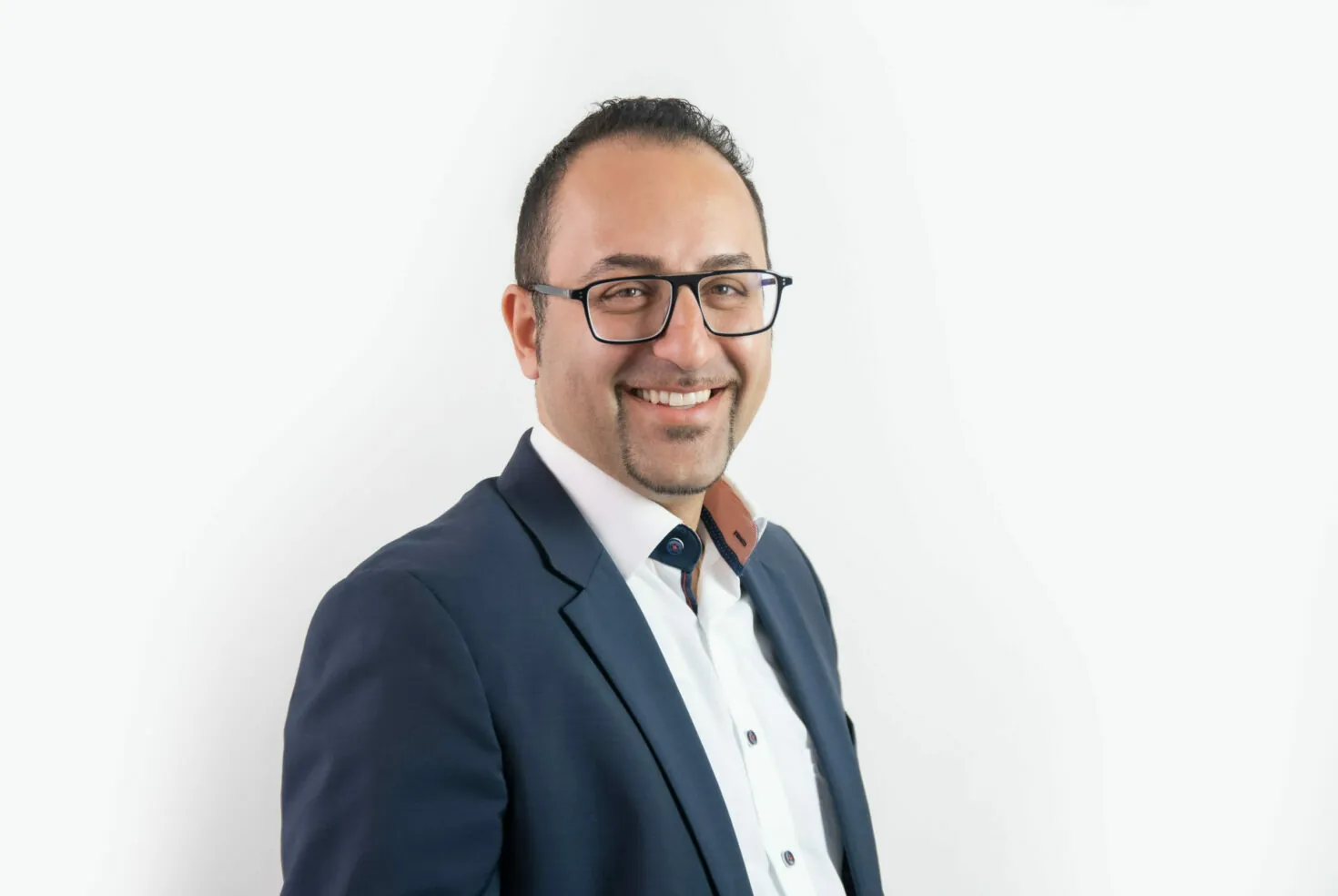 A man with glasses, wearing a white shirt and a dark blue blazer, smiles while standing against a white background.
