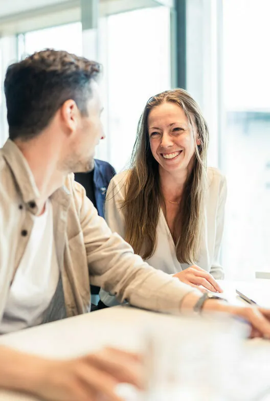 Two people sitting at a table, engaged in conversation and smiling. One person is wearing a beige jacket and a white shirt, while the other wears a white top. Background shows blurred windows.