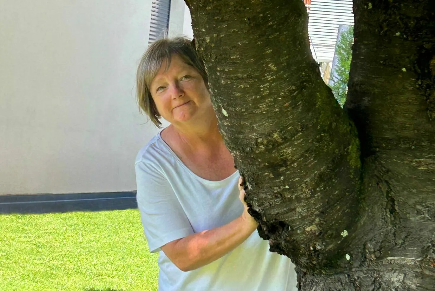 A woman in a light-colored shirt stands behind a tree trunk, with a portion of a building and grassy area in the background.