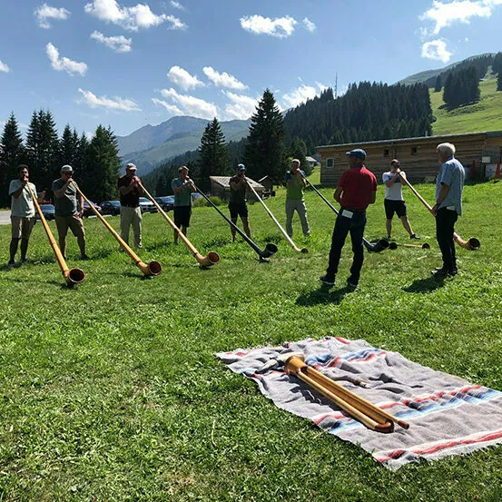 A group of people, standing in a semi-circle on a grassy field, playing long wooden alphorns in a mountainous area on a sunny day. A blanket with additional alphorns lies on the grass in the foreground.