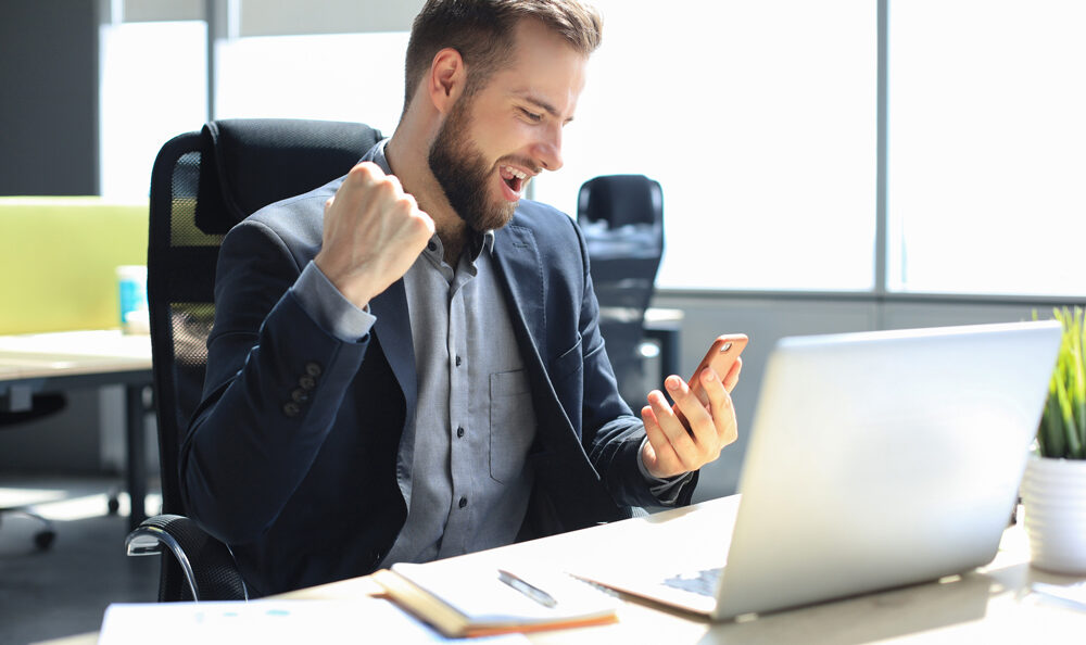 Mann im Anzug sitzt an einem Schreibtisch, blickt mit erhobener Faust auf sein Smartphone, in einer hell erleuchteten Büroumgebung. Auf dem Schreibtisch stehen ein Laptop, ein Notizbuch und eine Topfpflanze.