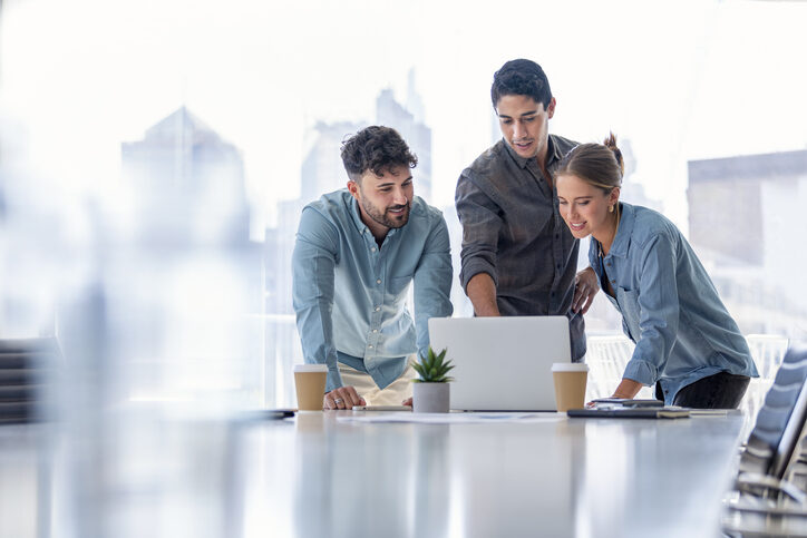 Business-Team, das an einem Laptop-Computer arbeitet. Drei Personen tragen Freizeitkleidung. Sie stehen in einem Sitzungssaal
