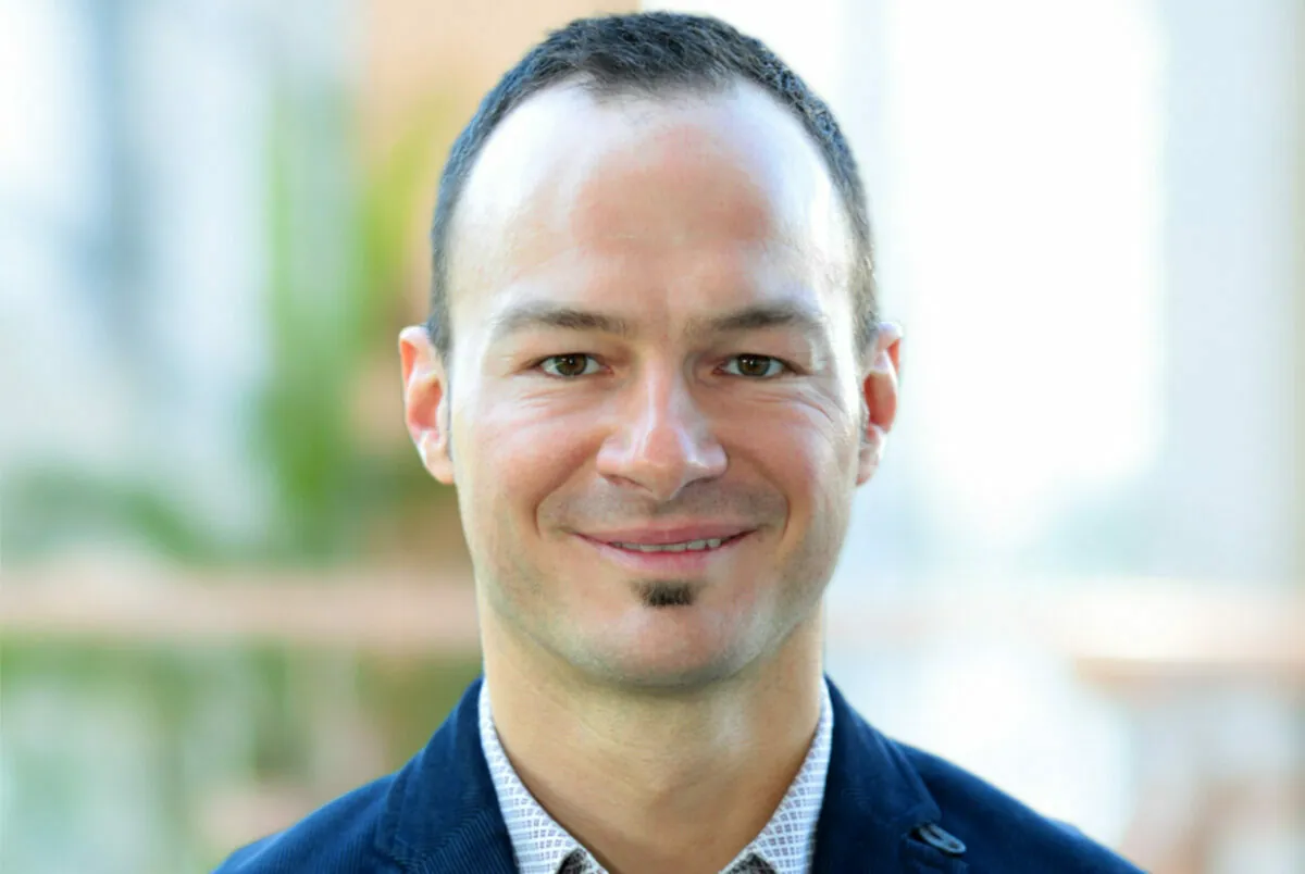A man with short dark hair and a small goatee is smiling. He is wearing a dark blue jacket and a patterned shirt. The background is softly blurred.