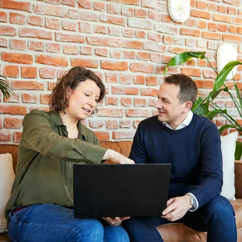 A woman and a man sit on a couch in a modern office, looking at a laptop on the woman's lap. The brick wall and green plants in the background add to the contemporary setting.
