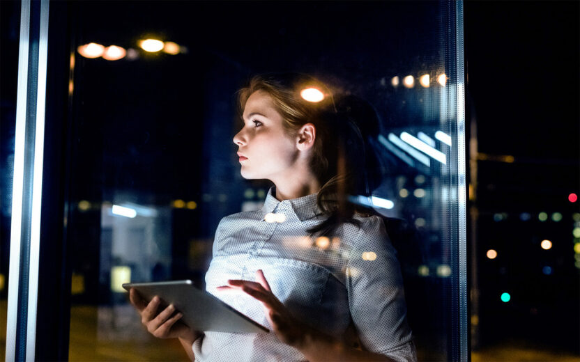Young businesswoman with tablet in the office working late at night. Shot through glass.