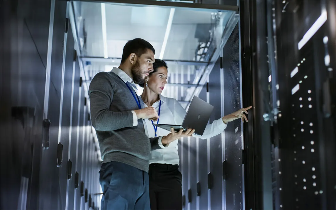 Male IT Specialist Holds Laptop and Discusses Work with Female Server Technician. They're Standing in Data Center, Rack Server Cabinet is Open. | Business Analytics