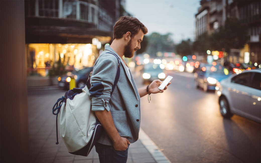 Young man on the street of big city , waiting taxi, chacking his smartphone, for news or new messages. Or looking for map instructions. Carry backpack on one shoulder. Dusk time. Casual dressed. Street lights in background.