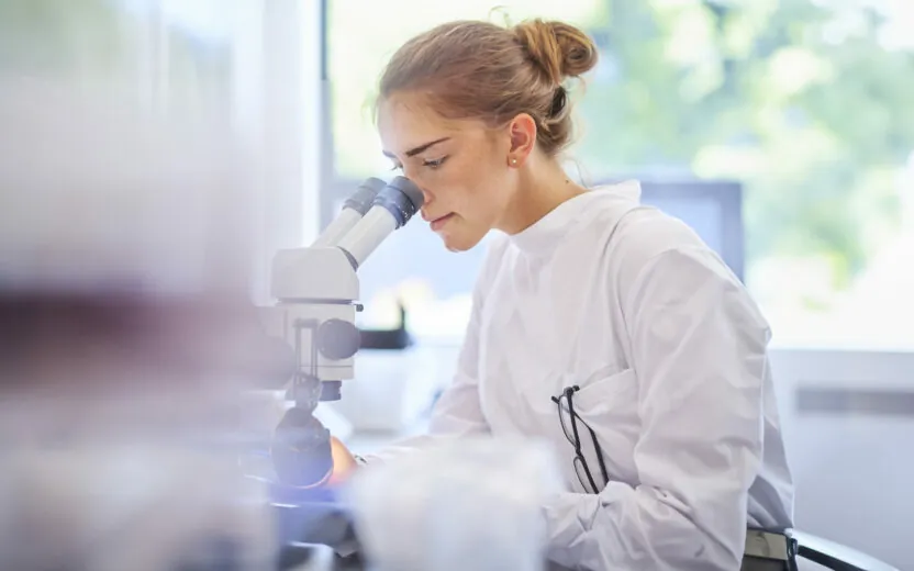 A female research scientist is analysing a sample on her microscope in a microbiology lab . The lab is brightly lit with natural light. Blurred glassware at side of frame provides copy space