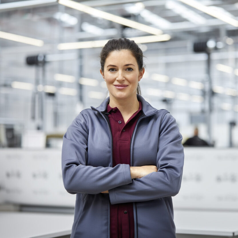 Portrait of female engineer with arms crossed in automobile industry. Confident technologist is standing in showroom. She is in jacket at factory.