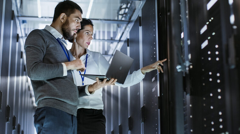 Männlicher IT-Spezialist hält Laptop und bespricht Arbeit mit weiblichem Servertechniker. Sie stehen im Rechenzentrum, der Serverschrank ist geöffnet.