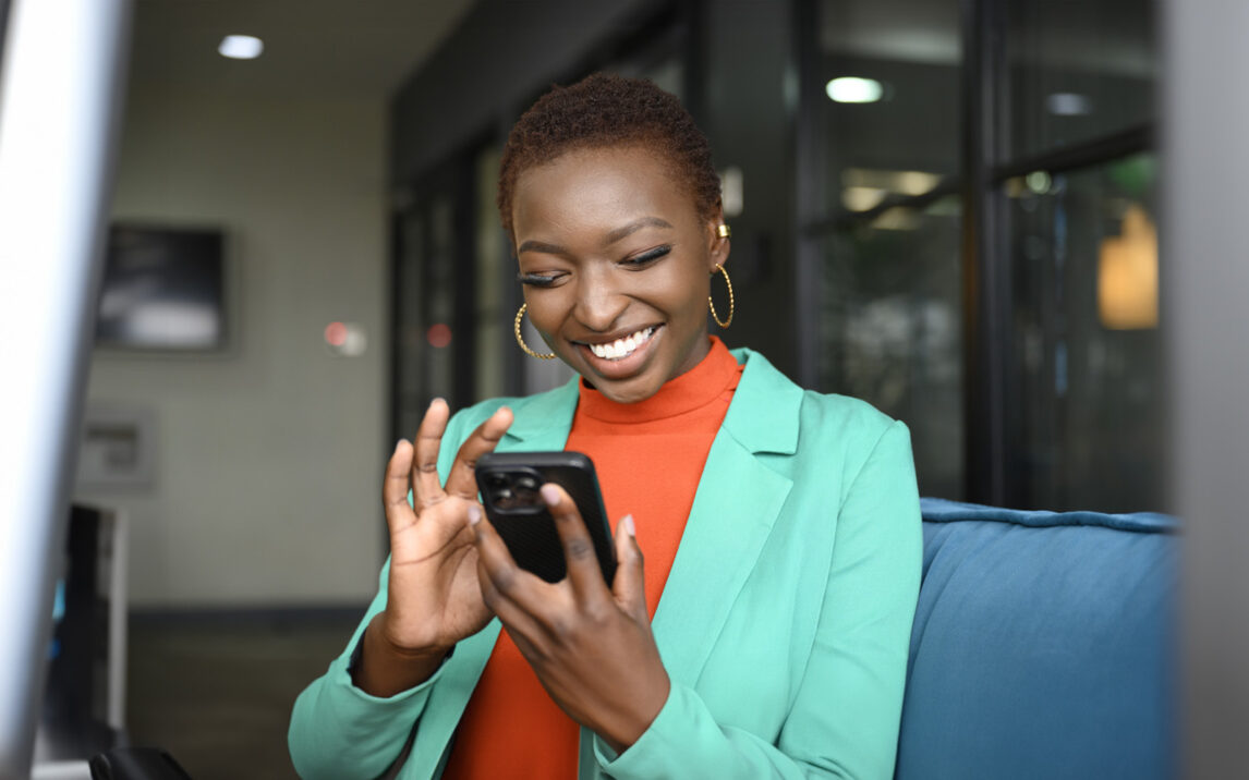 Waist-up view of shorthaired woman wearing hoop earrings, teal jacket over orange top, and smiling while scrolling portable device in modern office.