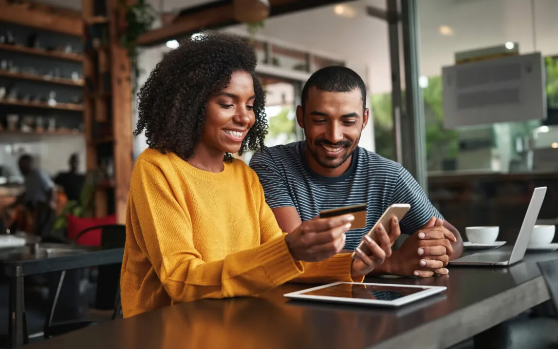 Young man looking at his friend using credit card and smartphone for shopping online in cafe