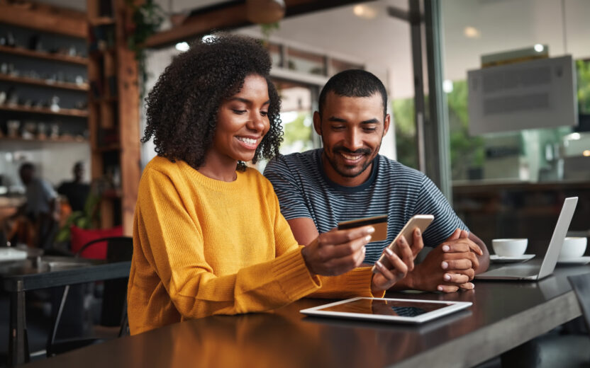 Young man looking at his friend using credit card and smartphone for shopping online in cafe