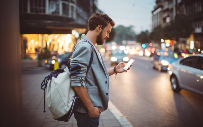Young man on the street of big city , waiting taxi, chacking his smartphone, for news or new messages. Or looking for map instructions. Carry backpack on one shoulder. Dusk time. Casual dressed. Street lights in background.