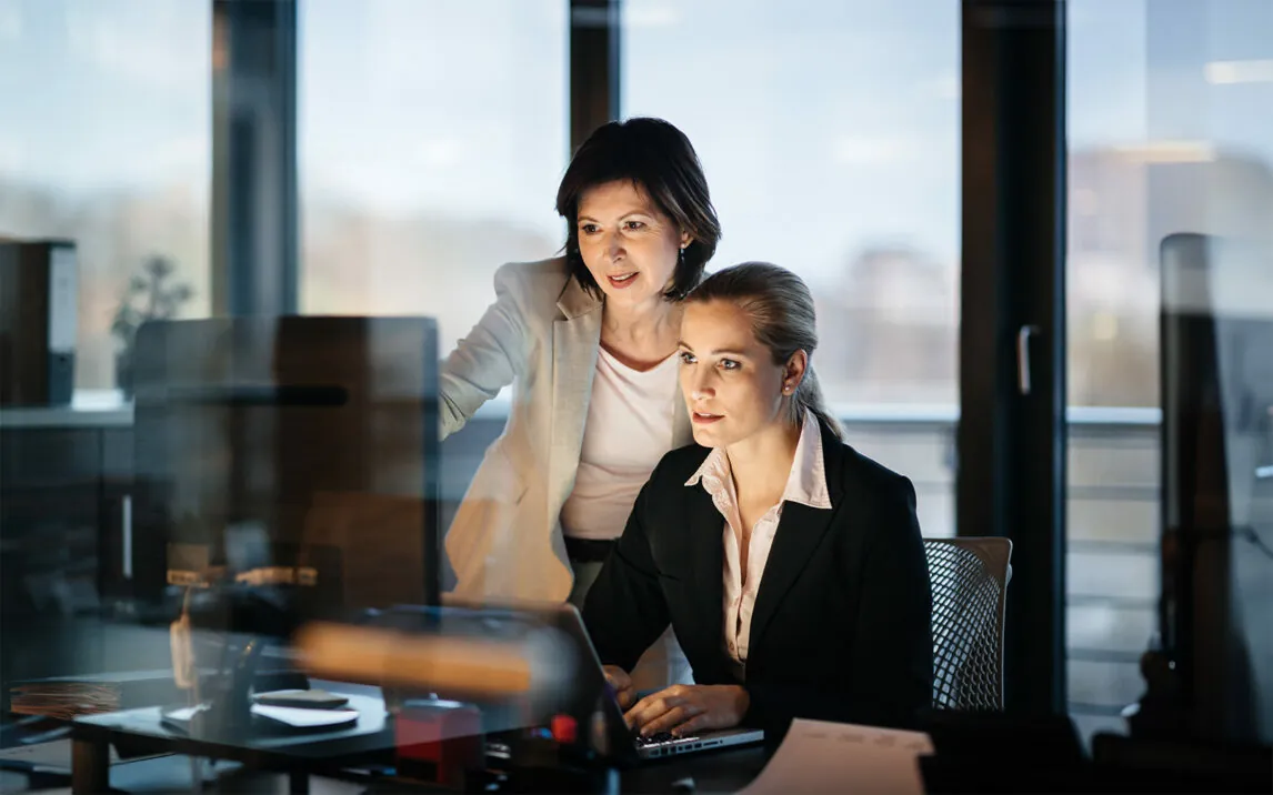 A young and mature businesswoman working late in front of a laptop and monitor in a modern office space. both faces are illuminated only by the computer monitor.