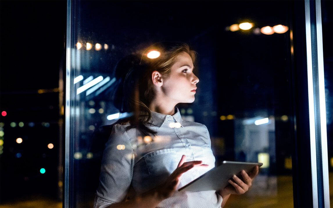 Young businesswoman with tablet in the office working late at night. Shot through glass.