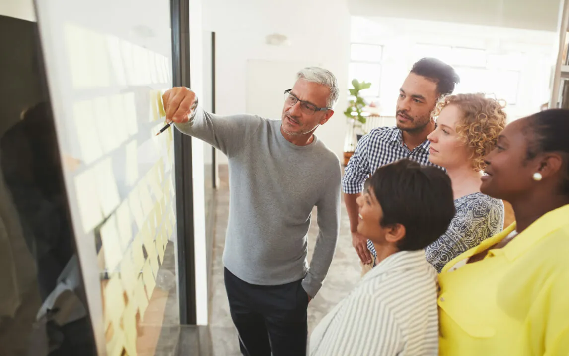 Mature businessman and his diverse team brainstorming with yellow adhesive notes on a glass wall in a bright modern office