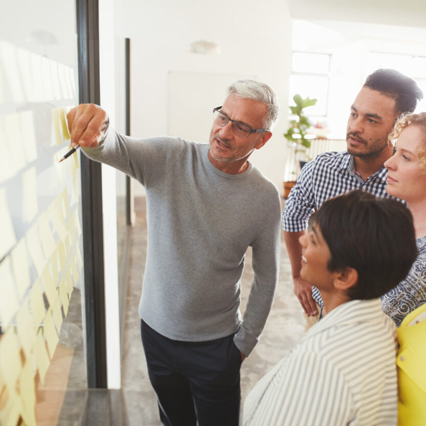 Mature businessman and his diverse team brainstorming with yellow adhesive notes on a glass wall in a bright modern office