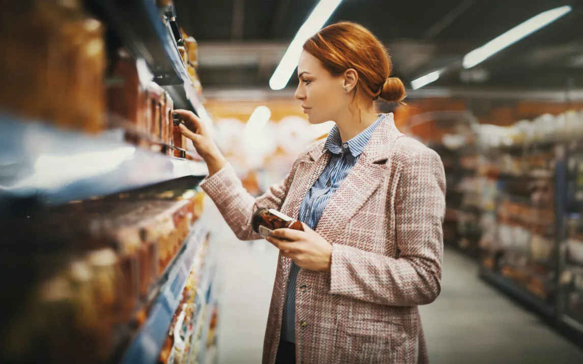 Young woman holding a jar of Sun-dried tomatoes in supermarket. She's searching for specific manufacturer.