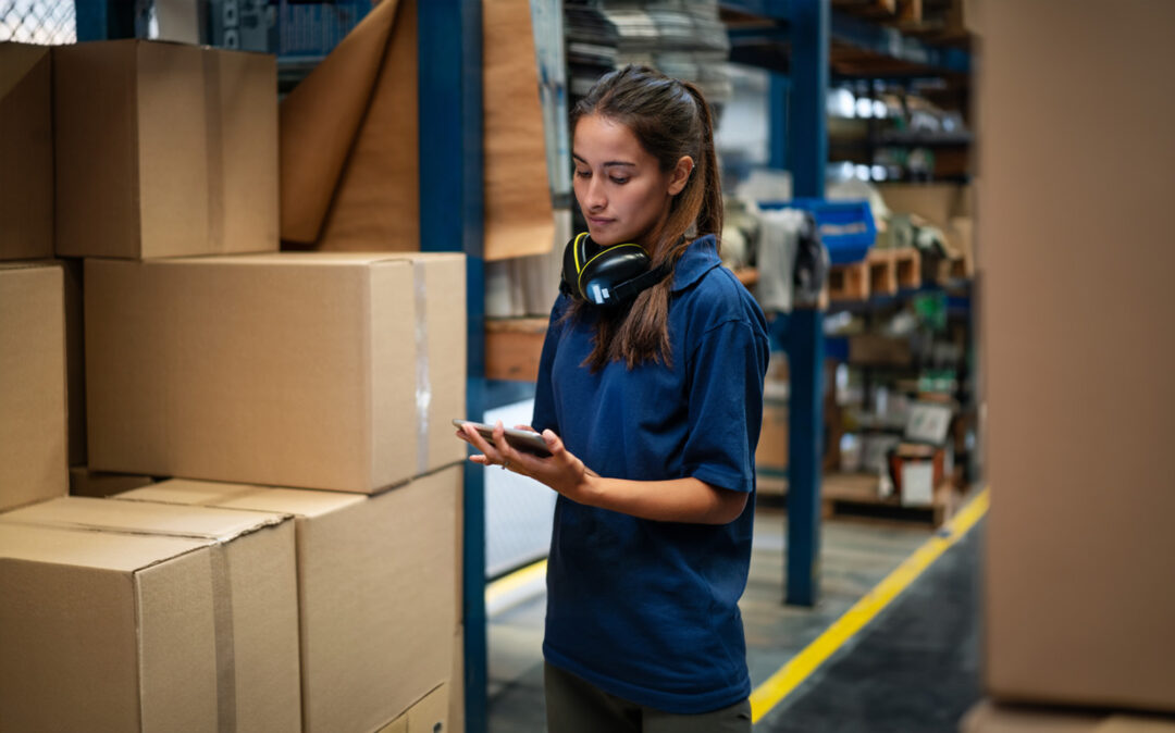 Female warehouse worker updating the stock on mobile phone app. Woman in uniform working in a factory warehouse.