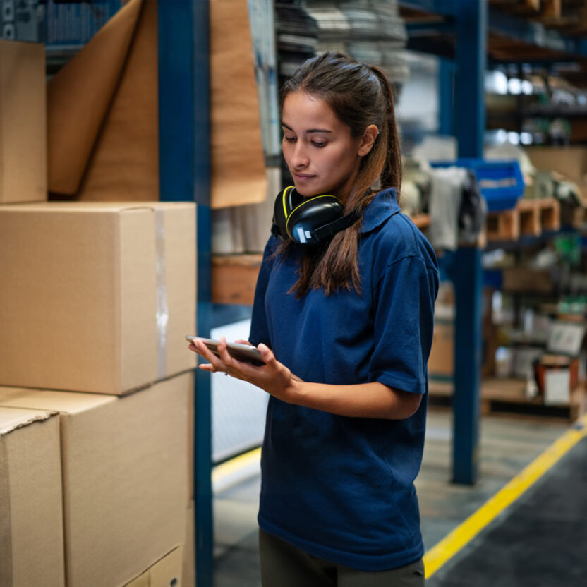 Female warehouse worker updating the stock on mobile phone app. Woman in uniform working in a factory warehouse.