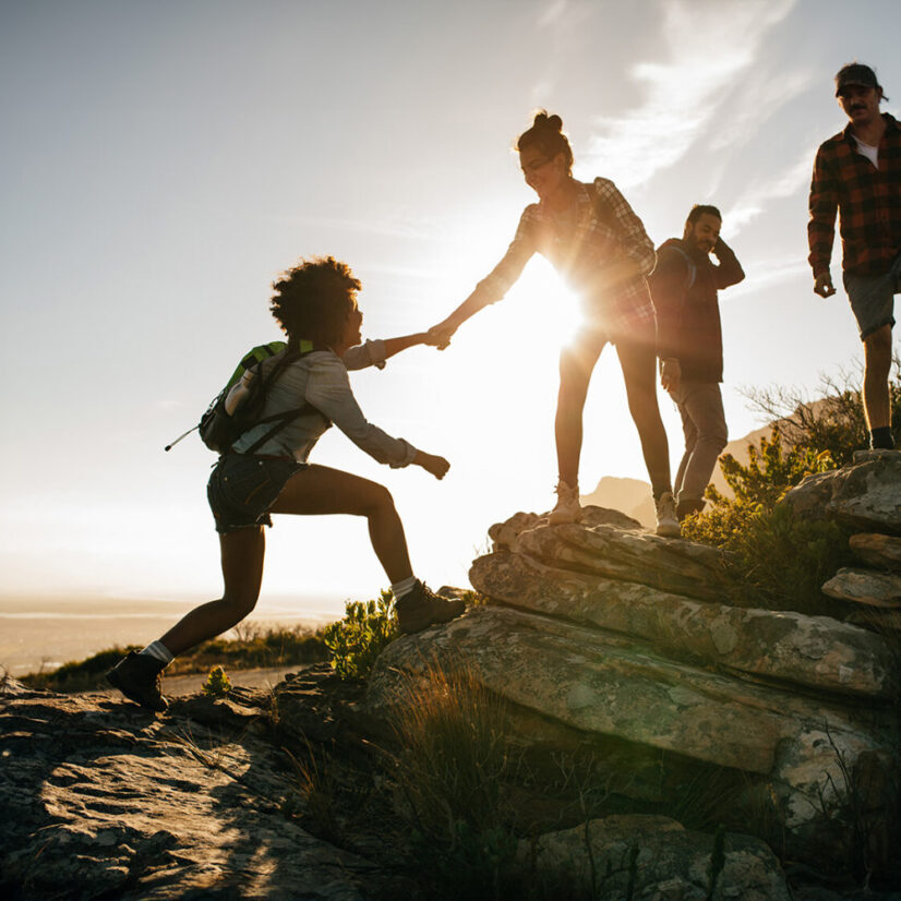 Group of hikers on a mountain. Woman helping her friend to climb a rock. Young people on mountain hike at sunset.